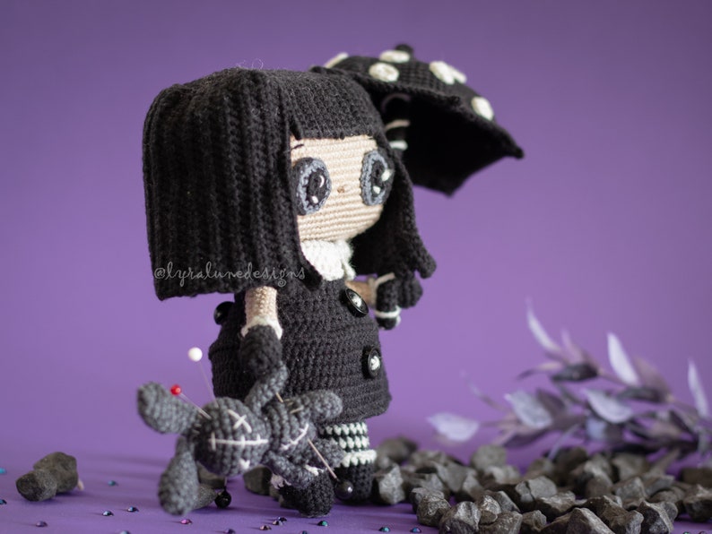 May include: A black crocheted doll with long black hair, wearing a black dress and a white collar. The doll is holding a small black crocheted doll with an X on its chest. The doll is standing on a purple surface with black rocks and glitter.
