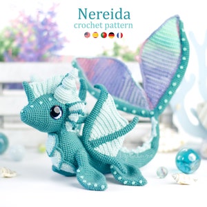 May include: A crocheted sea dragon amigurumi toy with a teal green body, white and light blue wings, and a light blue and purple tail. The dragon has a white horn and large blue eyes. The toy is sitting on a white surface with seashells and blue glass marbles.
