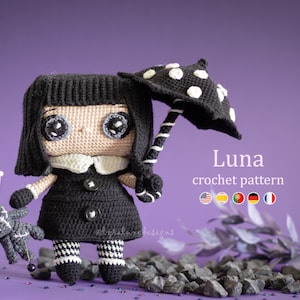 May include: A crocheted doll named Luna with black hair, a black dress, and white and black striped leggings. She is holding a black and white crocheted umbrella and a gray crocheted doll with a pin through it. The doll is standing on a bed of gray rocks.