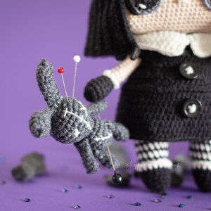 May include: A black and white crocheted doll with black buttons and a white collar holds a gray crocheted voodoo doll with red and white pins. The doll is surrounded by scattered black and silver glitter on a purple background.