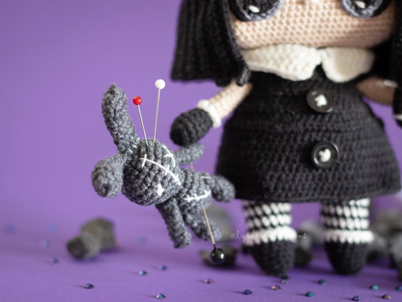 May include: A black and white crocheted doll with black buttons and a white collar holds a gray crocheted voodoo doll with red and white pins. The doll is surrounded by scattered black and silver glitter on a purple background.