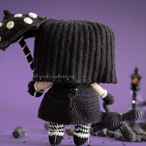 May include: A black crocheted doll wearing a black coat and a black and white polka dot umbrella. The doll is holding a black crocheted dog. The doll is standing on a purple surface with black and white confetti.