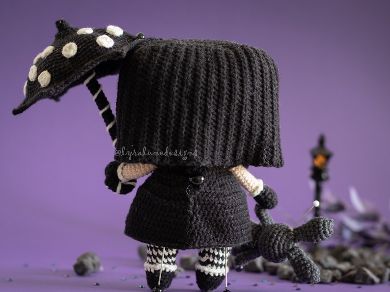May include: A black crocheted doll wearing a black coat and a black and white polka dot umbrella. The doll is holding a black crocheted dog. The doll is standing on a purple surface with black and white confetti.