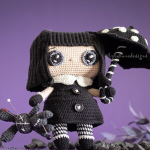 May include: A black crocheted doll with large eyes and a white collar holds a black and white polka dot umbrella. The doll is wearing a black dress with white and black checkered trim. A small black crocheted doll with pins is lying on the ground.