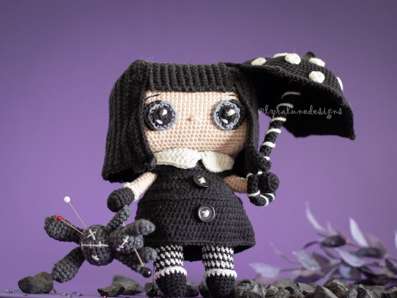 May include: A black crocheted doll with large eyes and a white collar holds a black and white polka dot umbrella. The doll is wearing a black dress with white and black checkered trim. A small black crocheted doll with pins is lying on the ground.