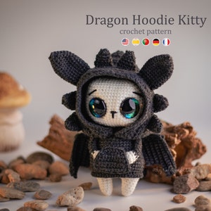 May include: A crocheted amigurumi kitty wearing a black dragon hoodie. The kitty has large, black eyes with green pupils and a white face. The hoodie has a black and gray color scheme and features a dragon-like hood with spikes.
