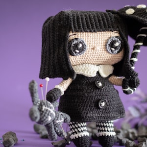 May include: A crocheted doll with black hair, a black dress, and white and black striped leggings. The doll is holding a black and white umbrella with white polka dots. The doll is standing on a purple surface with black rocks.