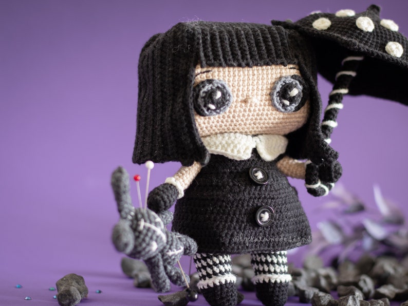 May include: A crocheted doll with black hair, a black dress, and white and black striped leggings. The doll is holding a black and white umbrella with white polka dots. The doll is standing on a purple surface with black rocks.