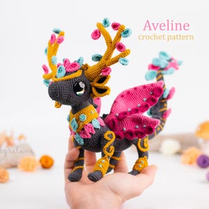 May include: A crocheted creature with a dark gray body, adorned with gold, pink, and turquoise accents. It has large, expressive eyes and decorative antlers. The text "Aveline crochet pattern" is visible.