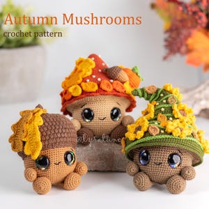 Op de afbeelding: Drie gehaakte paddenstoelenfiguren met grote, expressieve ogen en lachende gezichten. Eén is een eikel en de andere zijn paddenstoelen met kleurrijke doppen. De tekst "Autumn Mushrooms crochet pattern" staat bovenaan. De figuren zijn bruin, oranje en geel.