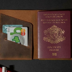 May include: Open brown leather passport holder with a burgundy passport and two credit cards. The passport has gold lettering and a coat of arms. The credit cards are green and white.