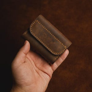 May include: A brown leather wallet with a curved flap, held in a hand. The wallet has visible stitching in a contrasting yellow-brown thread. The leather has a distressed appearance, with variations in color.
