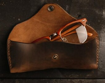 May include: A brown leather glasses case with a snap closure, open to reveal a pair of orange-framed spectacles. The case is made of distressed leather and has a stitched edge.