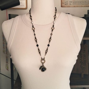May include: A necklace featuring a bronze-toned chain with intricate circular links, interspersed with small white pearls and black beads. The chain terminates in a central pendant consisting of a dark, spherical stone held by a bronze claw-like setting.