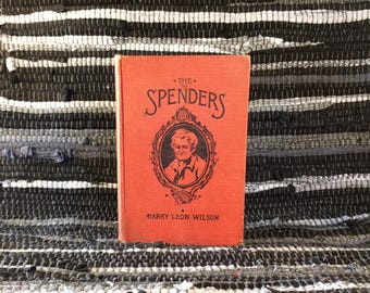 May include: A vintage book titled "The Spenders" by Harry Leon Wilson. The book has a red cover with black lettering and an illustration of a man in a circular frame. The book is displayed on a black and white striped rug.