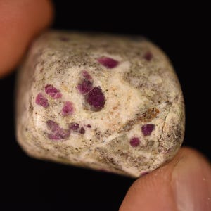 以下が含まれることがあります： 白と灰色の岩のクローズアップ。小さな濃いピンク色のルビーのインクルージョンがあります。