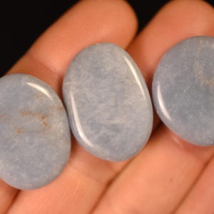 May include: Three smooth, polished, oval-shaped stones with a pale, dusty blue color. Each stone features a glossy, reflective surface with subtle, cloudy white and light brown internal inclusions visible throughout the light blue mineral structure.