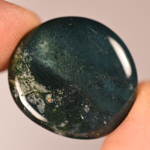 May include: A polished, oval-shaped moss agate gemstone. The stone features a deep green base color with intricate patterns of lighter green and white inclusions, creating a natural, organic appearance. The stone is held between two fingers.