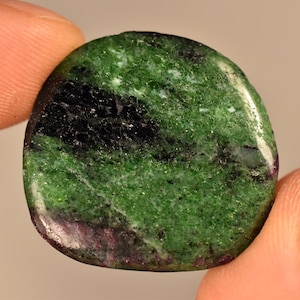 Może przedstawiać: Zielony, czarny i fioletowy kamień zoisitu o gładkiej, wypolerowanej powierzchni. Kamień ma owalny kształt i marmurkowy wygląd.