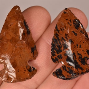 May include: Two obsidian arrowheads, each with a triangular shape and a notched base. The arrowheads display a warm brown hue with striking black markings. They are likely crafted for decorative or display purposes.