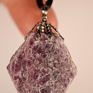 Puede incluir: Un colgante de rubí en bruto. La gema de color magenta oscuro está sujeta por un engaste de metal plateado y cuelga de un cordón negro. El rubí tiene una superficie rugosa y texturizada con variaciones naturales de color y tono.