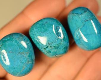 Könnte beinhalten: Drei polierte, getrommelte Türkissteine in leuchtendem Cyanblau. Jeder Stein hat eine glatte, glänzende Oberfläche mit natürlichen dunklen Aderungsmustern, die sich durch die Mineralstruktur ziehen und eine abgerundete, organische Form aufweisen.