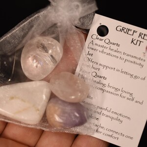 Könnte beinhalten: Ein Set Kristalle in einem weißen Organzasäckchen. Die Kristalle sind klarer Quarz, Jet, Rosenquarz und Amethyst. Die Tasche hat ein Etikett mit der Aufschrift "Trauerhilfe-Set" und listet die Eigenschaften jedes Kristalls auf.