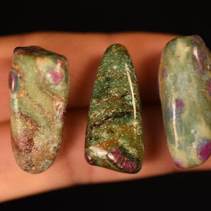 May include: Three polished tumbled stones featuring a mix of green, tan, and brown mineral patterns with embedded vibrant magenta-colored inclusions. The stones have a smooth, glossy surface texture and irregular, rounded shapes.