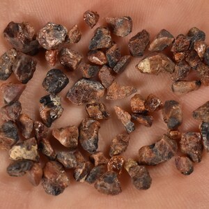 Puede incluir: Una colección de pequeñas piedras preciosas ásperas de color marrón y negro. Las piedras preciosas tienen una forma irregular y un ligero brillo metálico.