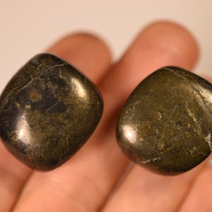 May include: Two polished, rounded stones with a metallic, dark olive-green and bronze-toned surface. The stones have a smooth, reflective texture with visible natural mineral inclusions and irregular dark patches across their rounded forms.