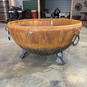 May include: A large, round, bowl-shaped fire pit made of rusted metal with a dark, charred-looking base. It features two hexagonal metal handles on the sides and stands on three curved, dark gray metal legs. A small oval metal logo plate is visible.