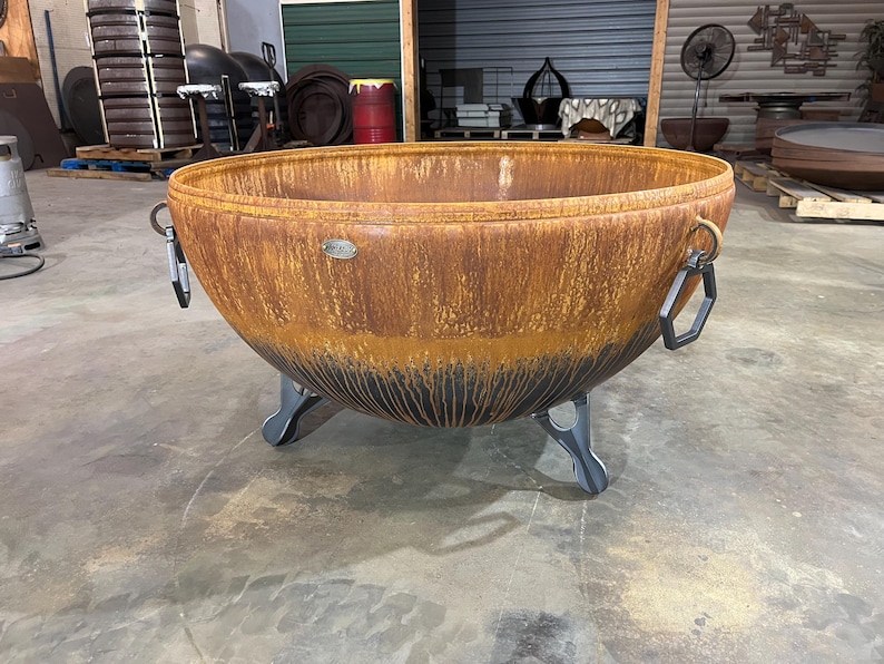 May include: A large, round, bowl-shaped fire pit made of rusted metal with a dark, charred-looking base. It features two hexagonal metal handles on the sides and stands on three curved, dark gray metal legs. A small oval metal logo plate is visible.