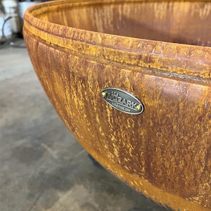 May include: A large, bowl-shaped fire pit with a weathered, rusted orange and brown patina finish. An oval-shaped metal plaque is attached to the exterior rim, featuring the embossed text MOZARK and a small graphic icon above the brand name.