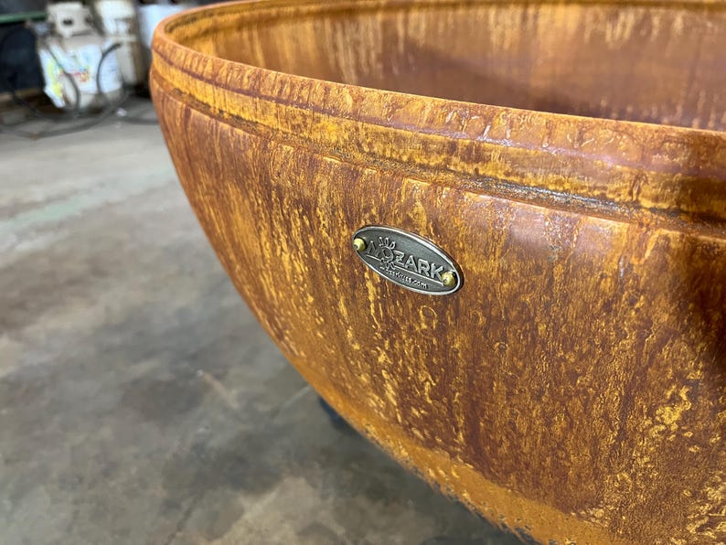 May include: A large, bowl-shaped fire pit with a weathered, rusted orange and brown patina finish. An oval-shaped metal plaque is attached to the exterior rim, featuring the embossed text MOZARK and a small graphic icon above the brand name.