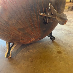 May include: A large, spherical metal fire pit with a dark, oxidized finish featuring prominent rust-colored streaks radiating from the center. The bowl is supported by three sturdy, curved metal legs and includes a welded side handle attachment.