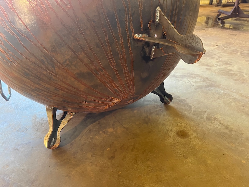 May include: A large, spherical metal fire pit with a dark, oxidized finish featuring prominent rust-colored streaks radiating from the center. The bowl is supported by three sturdy, curved metal legs and includes a welded side handle attachment.