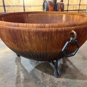 May include: A large, hemispherical metal fire pit with a weathered, rusted orange and brown patina. The bowl features a thick, rolled rim, a black hexagonal handle attached to a metal ring on the side, and three sturdy, dark gray metal legs.