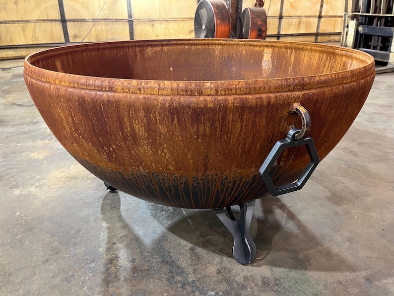 May include: A large, hemispherical metal fire pit with a weathered, rusted orange and brown patina. The bowl features a thick, rolled rim, a black hexagonal handle attached to a metal ring on the side, and three sturdy, dark gray metal legs.