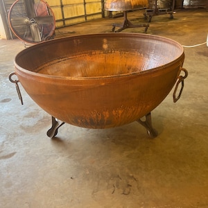 May include: A large, round, rust-colored metal fire pit bowl with a weathered patina. The bowl features three sturdy, curved metal legs for support and two hexagonal metal ring handles attached to opposite sides of the exterior rim.