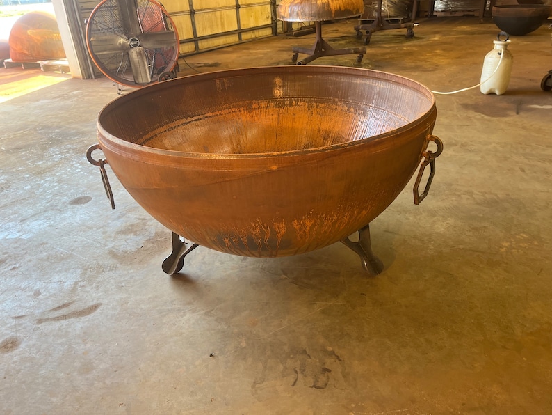 May include: A large, round, rust-colored metal fire pit bowl with a weathered patina. The bowl features three sturdy, curved metal legs for support and two hexagonal metal ring handles attached to opposite sides of the exterior rim.