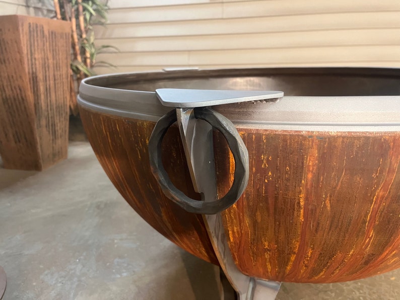 Puede incluir: Una gran hoguera de metal en forma de cuenco con un acabado exterior oxidado de color marr&oacute;n anaranjado. El borde presenta un soporte met&aacute;lico triangular gris liso unido a un asa de anillo circular forjada a mano, oscura y texturizada.