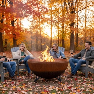 May include: A large, round, rust-colored metal fire pit with a textured exterior and black metal legs, containing a bright, active flame. The fire pit is positioned outdoors in a wooded area covered in fallen autumn leaves during the golden hour.