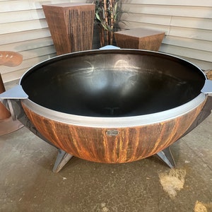 Puede incluir: Una hoguera grande y redonda en forma de cuenco con un acabado exterior de color naranja y marr&oacute;n oxidado. El interior es de metal negro liso. Cuenta con tres patas de metal resistentes, un borde plateado y tres acentos triangulares plateados.