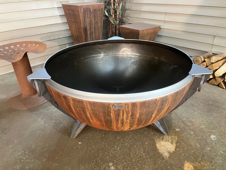 Puede incluir: Una hoguera grande y redonda en forma de cuenco con un acabado exterior de color naranja y marr&oacute;n oxidado. El interior es de metal negro liso. Cuenta con tres patas de metal resistentes, un borde plateado y tres acentos triangulares plateados.