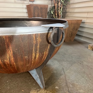 Puede incluir: Una gran hoguera en forma de cuenco hecha de metal oxidado y desgastado con una p&aacute;tina naranja y marr&oacute;n. Presenta un borde de metal gris liso, patas triangulares resistentes y un asa de anillo de metal circular unida al lateral para moverla.