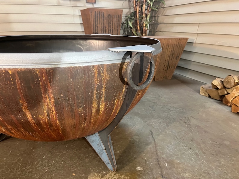 Puede incluir: Una gran hoguera en forma de cuenco hecha de metal oxidado y desgastado con una p&aacute;tina naranja y marr&oacute;n. Presenta un borde de metal gris liso, patas triangulares resistentes y un asa de anillo de metal circular unida al lateral para moverla.