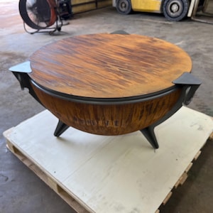 Puede incluir: Un brasero redondo con un acabado de metal oxidado y desgastado en el cuenco exterior y una tapa circular a juego. La estructura se apoya en tres patas met&aacute;licas oscuras y angulares, con detalles decorativos que sobresalen del borde.