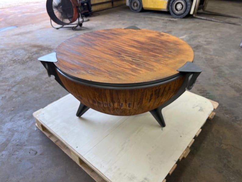 Puede incluir: Un brasero redondo con un acabado de metal oxidado y desgastado en el cuenco exterior y una tapa circular a juego. La estructura se apoya en tres patas met&aacute;licas oscuras y angulares, con detalles decorativos que sobresalen del borde.