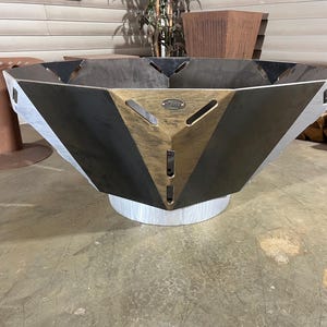 May include: A large, geometric fire pit with a metallic finish. The fire pit has a multi-faceted design with a black and gold center and silver sides. It sits on a silver base. The fire pit is designed for outdoor use.