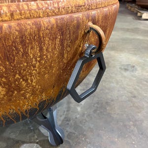 May include: A large, bowl-shaped fire pit with a weathered, rusted orange and brown patina. It features a matte black hexagonal metal handle attached to a circular ring on the side and is supported by a sturdy, dark gray metal tripod base.